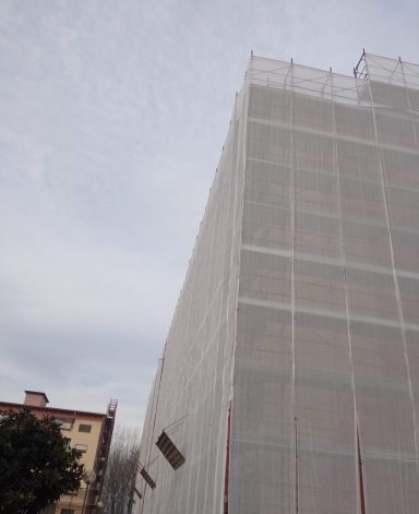 Edificio in ristrutturazione, coperto da una rete, con cielo nuvoloso sullo sfondo.